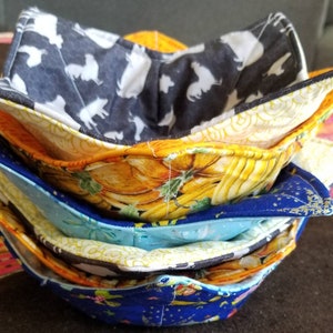 May include: A stack of four colorful fabric bowl cozies. The top cozy is black with white silhouettes of animals, the second is yellow with a pumpkin pattern, the third is blue with a star pattern, and the bottom is blue with a floral pattern.