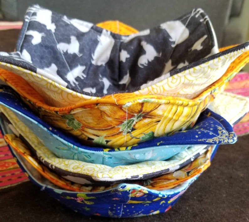 May include: A stack of four colorful fabric bowl cozies. The top cozy is black with white silhouettes of animals, the second is yellow with a pumpkin pattern, the third is blue with a star pattern, and the bottom is blue with a floral pattern.