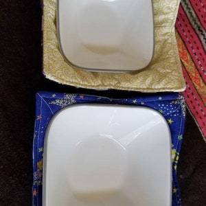 May include: Two white ceramic bowls with gray rims, one in a yellow and white patterned bowl cozy and the other in a blue and white patterned bowl cozy.