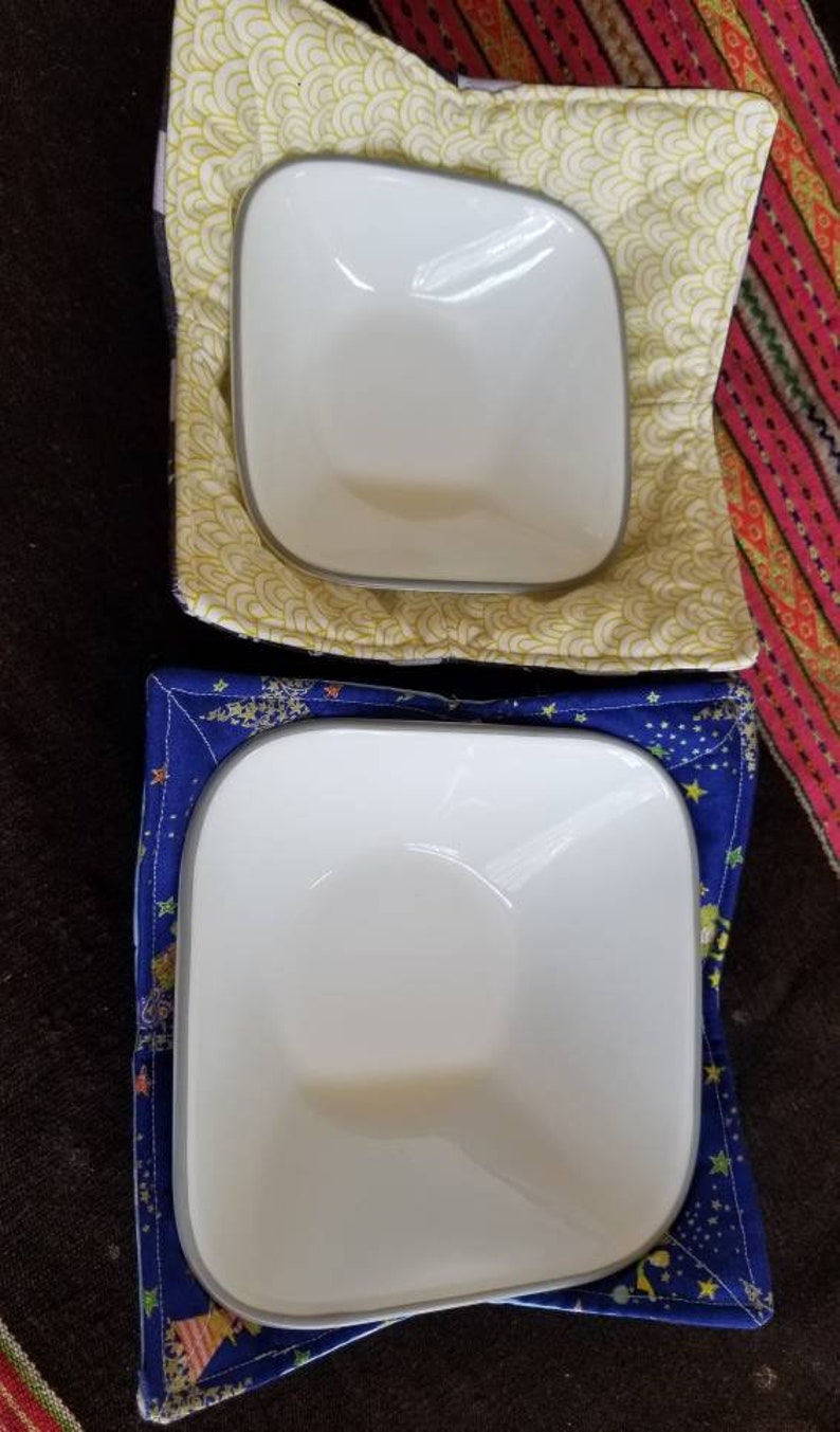 May include: Two white ceramic bowls with gray rims, one in a yellow and white patterned bowl cozy and the other in a blue and white patterned bowl cozy.