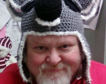 May include: A person wearing a gray and white crocheted hat shaped like a koala. The hat has large ears and a black nose.