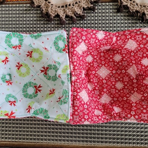 May include: Two fabric bowl cozies. One is white with green and red wreaths, the other is red with white and pink paisley designs.