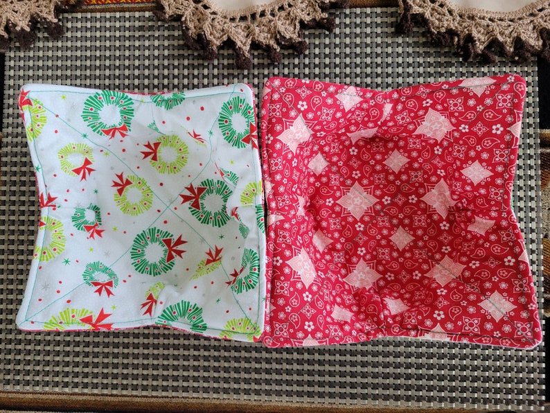 May include: Two fabric bowl cozies. One is white with green and red wreaths, the other is red with white and pink paisley designs.