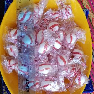 May include: A yellow bowl filled with individually wrapped peppermint candies. The candies are white with red stripes and are wrapped in clear plastic with a red and white snowflake design. The text "Sweet Stripes" is printed on the wrappers.