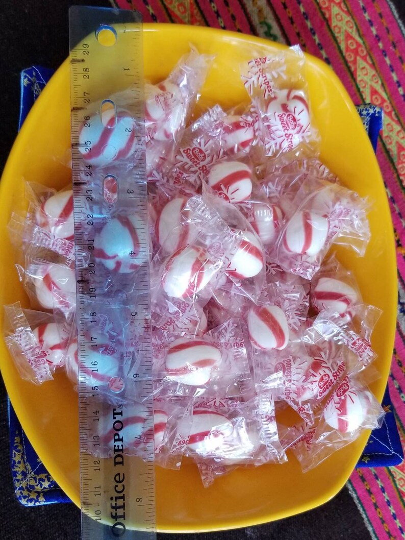 May include: A yellow bowl filled with individually wrapped peppermint candies. The candies are white with red stripes and are wrapped in clear plastic with a red and white snowflake design. The text "Sweet Stripes" is printed on the wrappers.