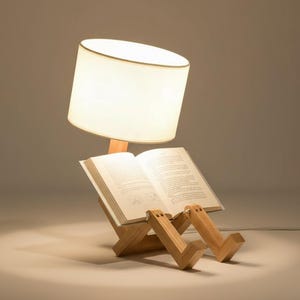 Könnte beinhalten: Ein Buchständer aus hellem Holz in Form einer sitzenden Figur mit beweglichen Beinen und Armen, die ein offenes Buch halten. Ein zylindrischer weißer Lampenschirm ist an der Holzstruktur befestigt und wirft ein warmes Licht auf die Buchseiten.