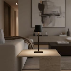 Pode incluir: Uma lâmpada de mesa preta com base retangular e haste fina, encimada por uma abajur preto. A lâmpada está colocada sobre uma mesa quadrada de cor clara em uma sala de estar. O fundo inclui um sofá, obras de arte e uma mesa de centro escura.
