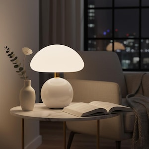 May include: A white mushroom-shaped table lamp with a gold base sits on a marble-topped side table. A white vase with a sprig of greenery and an open book are also on the table. The background shows a window with a city view.