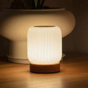 May include: A glowing white LED lamp with a ribbed, cylindrical shade sits on a round wooden base. The lamp is turned on, emitting a soft, warm light. The background features a large white pot and a wooden surface.