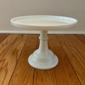 May include: A white milk glass cake stand featuring a smooth, circular flat top surface with a raised edge, supported by a central pedestal base that flares out at the bottom. The stand has a glossy, opaque finish and a clean, simple design.