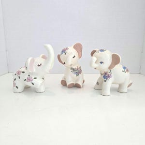 Puede incluir: Tres figuras de elefantes de cerámica blanca con motivos florales pintados. El elefante de la izquierda tiene flores rosas, el del centro tiene flores azules y moradas con una etiqueta que dice HAPPY, y el de la derecha tiene flores azules y moradas.