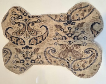 May include: A bone-shaped textile mat featuring a beige background with a repeating dark blue and brown paisley and floral pattern. The edges are finished with a stitched border, creating a distinct shape with four rounded corners.