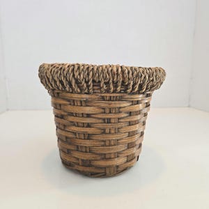 May include: A small, round, woven basket with a natural brown color. The basket has a woven rim and a sturdy, textured body. This decorative basket is suitable for home decor and storage.