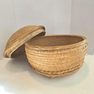 May include: A round, woven basket with a matching lid. The basket is made of light brown wicker and has a textured surface. The lid is slightly ajar, revealing the basket's interior. This decorative basket is suitable for storage.