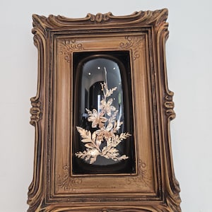 May include: A framed decorative piece featuring a black background with gold-colored floral accents inside a glass dome. The frame is ornate, with a gold-toned finish and detailed carvings, creating a vintage aesthetic.