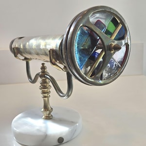 May include: A metallic kaleidoscope mounted on a brass stand with a white marble base. The viewing end features a circular frame with three spokes, containing segments of blue, green, and red stained glass that catch and reflect light.