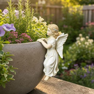 May include: A cream-colored angel figurine with detailed wings and a flowing dress, designed to hang over the edge of a large, textured, round gray planter filled with green foliage and small purple and white flowers in a garden setting.