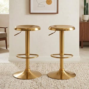 May include: Two gold-colored adjustable bar stools with round seats and footrests. The stools have a pedestal base and are set on a neutral-colored rug. A framed abstract art piece hangs on the wall behind the stools, and a wooden cabinet with plants is visible.