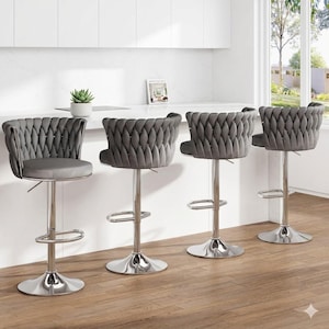May include: Four grey velvet bar stools with chrome bases and adjustable height. The stools feature a quilted back design and a footrest. They are set in a kitchen with a white countertop and wooden flooring. Each stool has a circular base.