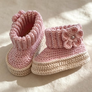 Pode incluir: Um par de sapatinhos de crochê rosa claro com punho canelado e sola bege. Um dos sapatinhos é adornado com uma flor de crochê na lateral, que inclui uma pequena conta prateada no centro. Os sapatinhos apresentam um padrão de ponto texturizado na parte superior e uma base bege sólida.