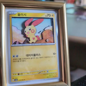 Könnte beinhalten: Eine gerahmte Pokémon-Karte mit einer gelb-roten Cartoon-Figur, dem Text "플러시" und "70 4". Die Karte befindet sich in einem goldfarbenen Rahmen. Die Karte hat koreanischen Text und Illustrationen.