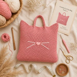 May include: A pink crocheted cat-shaped tote bag with two handles, featuring a small heart-shaped nose and white embroidered whiskers. The bag is displayed alongside a pattern card, yarn balls in a basket, crochet hooks, scissors, and a tape measure.