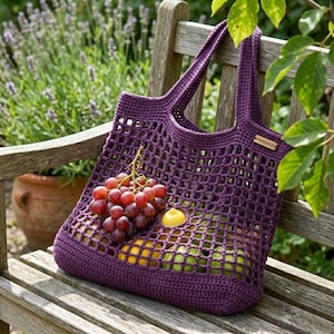 May include: A handmade, deep purple crochet tote bag filled with fresh fruit, including red grapes, lemons, and green apples. The bag is sitting on a wooden bench in a garden setting. The bag has a tag that says "HANDMADE".