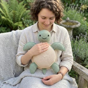 Peut inclure: Une peluche tortue crochetée, aux couleurs vert et beige. La tortue a un visage souriant et est tenue. La peluche a une carapace ronde et des appendices en forme de nageoires. Le fond présente de la verdure.