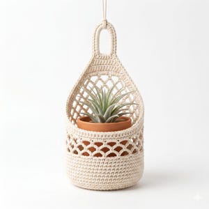 Op de afbeelding: Een crèmekleurige macramé plantenbak met een terracotta pot met een groene luchtplant. De plantenbak heeft een geweven ontwerp en een lus om op te hangen. De pot is rond en zit in de macramé houder.