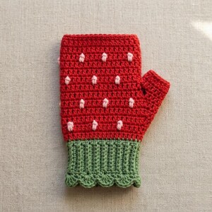 May include: A handmade crochet fingerless glove in the shape of a strawberry. The glove is primarily red with white and pink seed details. The cuff is green with a scalloped edge. The glove is designed to be worn on the left hand.