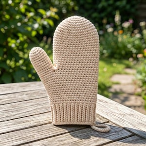 Op de afbeelding: Een beige gehaakte ovenwant met een ophanglus. De want is gemaakt van een getextureerd, gebreid materiaal. De duim is apart. De achtergrond toont een houten tafel en een groene tuin.