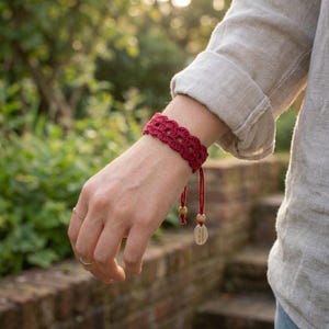 Può includere: Un braccialetto all'uncinetto rosso scuro caratterizzato da un motivo a bordo smerlato. Il braccialetto include due cordini pendenti, ciascuno rifinito con una piccola perla di legno e un piccolo ciondolo ovale in legno, per una chiusura regolabile.