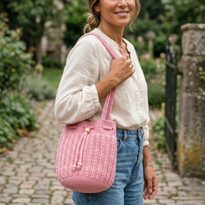May include: A pink crochet bucket bag with a drawstring closure and wooden bead accents. The bag has a textured pattern and a matching pink strap. The bag is held by a person wearing a white shirt and blue jeans.