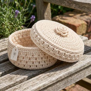 May include: A cream-colored, crocheted basket with a matching lid, resting on a wooden bench. The lid features a decorative flower and the basket has a tag with a flower illustration. The open basket reveals its interior, showcasing the intricate crochet pattern.