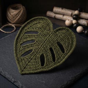 May include: A handmade, olive green crochet leaf-shaped coaster with three oval cutouts. The coaster is placed on a dark gray slate surface. A ball of twine and decorative wooden beads are in the background.