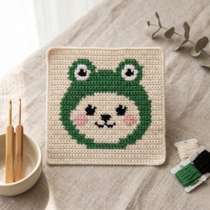 May include: A square, cream-colored crocheted potholder featuring a green frog face design. The frog has black eyes, pink cheeks, and a smiling mouth. Two wooden crochet hooks and a small bowl are visible.