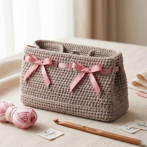 Op de afbeelding: Een gehaakte handtas in een neutrale beige kleur, versierd met roze strikken. De tas is rechthoekig met een textuursteekpatroon. Twee bollen roze en wit garen, een houten haaknaald en labels zijn ook aanwezig.