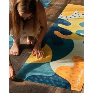 Pode incluir: Um tapete de ioga retangular com um padrão abstrato de formas orgânicas em tons de amarelo, laranja, azul-petróleo e azul-escuro. A superfície inclui texturas pontilhadas e o texto Downward Frog impresso em branco no canto inferior direito.