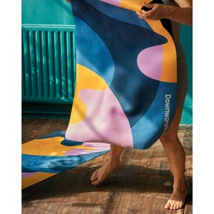Puede incluir: Una esterilla de yoga rectangular con un patrón abstracto de formas grandes y fluidas en azul marino, amarillo mostaza y rosa suave. El texto "Downward Frog" está impreso verticalmente en blanco a lo largo del borde derecho de la esterilla, parcialmente desenrollada.