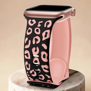 May include: Rose gold smartwatch with a black leopard print strap. The strap has a pink interior and a black leopard print design. The watch is displayed on a light-coloured, textured surface.