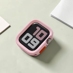 Pode incluir: Uma capa rosa para smartwatch com um ecrã preto que mostra as horas 10:09. A capa tem uma forma quadrada arredondada com um botão laranja na lateral. O relógio está numa superfície verde clara.