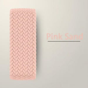 Puede incluir: Primer plano de una correa trenzada de color rosa pálido. La correa tiene un aspecto texturizado y tejido. Las palabras "Pink Sand" están impresas en rosa en el lado derecho de la imagen.