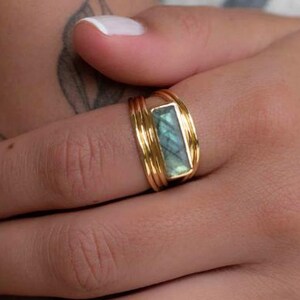 May include: A gold ring featuring a rectangular, iridescent green-grey labradorite gemstone set horizontally. The band is constructed from multiple thin, stacked gold wires that wrap around the finger, creating a layered, textured appearance.