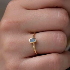 Puede incluir: Un anillo de oro con una pequeña piedra azul de corte rectangular engastada horizontalmente en una banda de oro fina y pulida. El anillo se lleva en un dedo, destacando el contraste entre el acabado metálico dorado y la piedra azul.