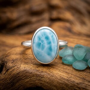 May include: A silver ring featuring a large, oval-shaped, light blue Larimar gemstone with white, cloud-like patterns. The stone is set in a smooth, polished silver bezel, and the ring band is made of a simple, polished silver metal.