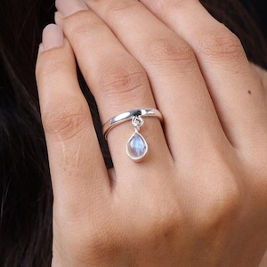 May include: A silver ring featuring a smooth, polished band and a single dangling charm. The charm is shaped like a teardrop and contains a small, iridescent moonstone that displays a soft blue glow against the metallic silver setting.