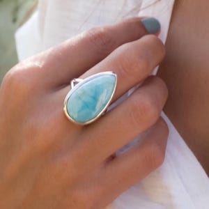 May include: A teardrop-shaped Larimar gemstone ring set in a polished silver band. The stone features a smooth, opaque surface with marbled patterns of sky blue and white, held securely by a simple metal bezel setting on a silver ring band.