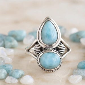 May include: A silver ring featuring two light blue, oval and teardrop-shaped stones set in a metal frame. The band splits into two sections, and the setting includes decorative metal detailing around the base of the stones with a rope-like texture.