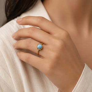 Könnte beinhalten: Ein goldener Ring mit einem runden, polierten hellblauen Stein in einer Fassung. Die Ringschiene weist eine texturierte, gehämmerte Oberfläche auf, die einen deutlichen Kontrast zu dem glatten, kreisförmigen Edelstein in der Mitte des Rings bildet.