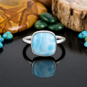 May include: A silver ring featuring a smooth, square-shaped, light blue Larimar gemstone with white marbled patterns. The ring has a polished metal band and is centered, displaying a clear reflection on the dark, glossy surface beneath it.